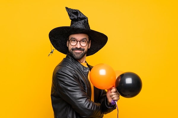 Happy Halloween man in a witch hat with glasses and a leather jacket holding orange and black balloons